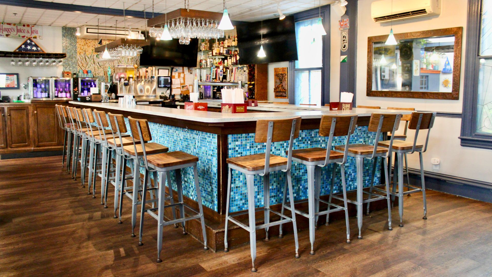 Bar area of a restaurant with a colorful mosaic tile wall, stools along bar countertop and overhead hanging gasses